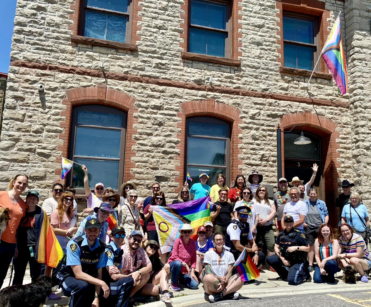 Un groupe de personnes est rassemblé à l’extérieur d’un bâtiment en pierre, en place pour prendre une photo. Un drapeau de la Fierté est brandi au milieu de la foule, et un autre flotte sur un mât au-dessus de l’entrée du bâtiment. Les gens portent eux-mêmes de plus petits drapeaux de la Fierté. Ils sourient à la caméra.