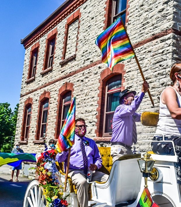 Deux personnes à bord d’une calèche blanche portent des chemises lavande et agitent des drapeaux de la Fierté. Au bord de la photo, on voit une autre personne à l’avant. La scène se passe devant un bâtiment en pierre. 
