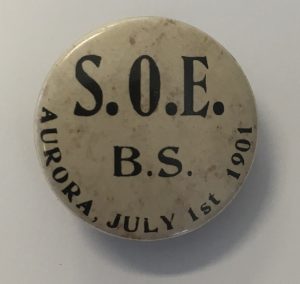 The front of a small circular button that has a tan/brown background with black text on it.