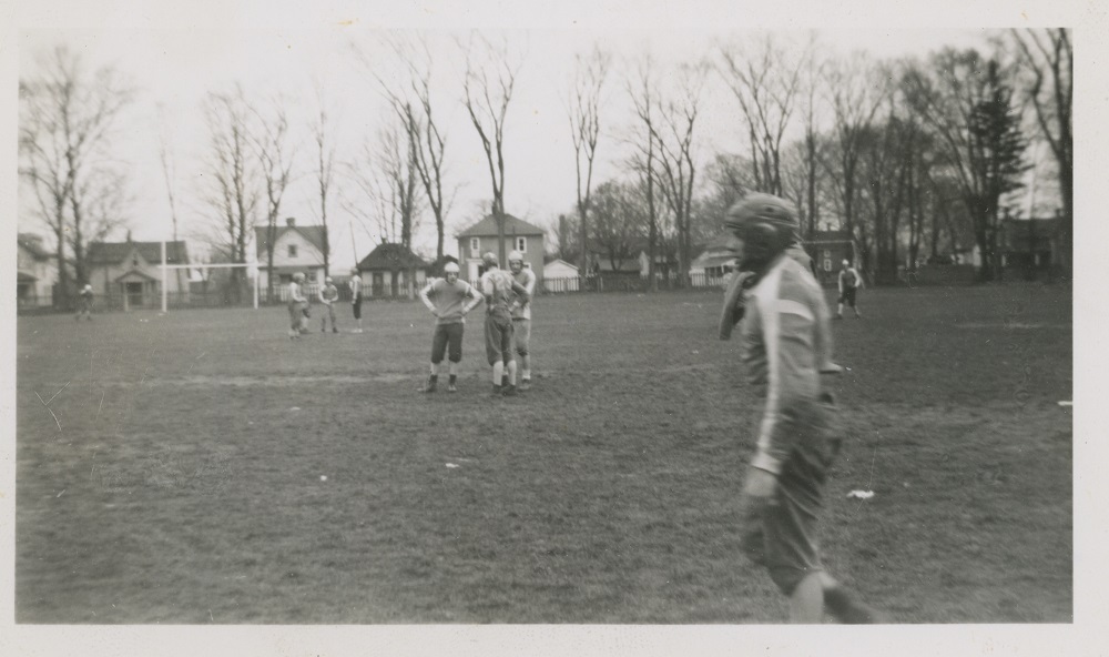 Photo en noir et blanc d’un groupe de jeunes joueurs de foot en uniforme sur une pelouse.  On voit des maisons en arrière plan derrière une barrière et une rangée d’arbres.