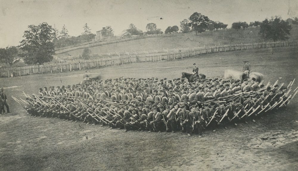 Photographie en noir et blanc de soldats en rangs serrés dans un champ, armés de fusils,  avec trois chevaux, deux assez flous, derrière deux hommes à gauche et au fond une ferme et une barrière en bois.