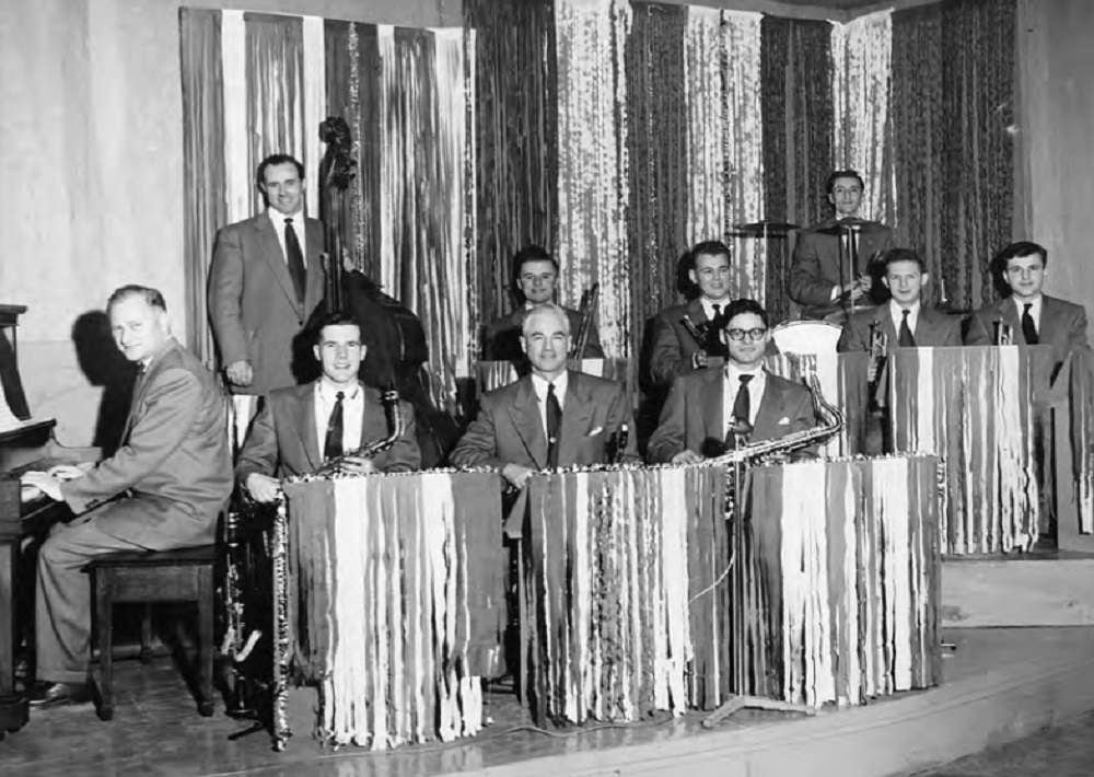 Photo en noir et blanc un peu floue d’un orchestre de dix musiciens en costume, cravate, derrière des pupitres sur une estrade à deux niveaux.  Des bandes verticales de papier ou de tissu donnent un effet chatoyant.  A gauche un pianiste derrière son instrument.