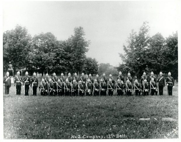 Photographie en noir et blanc de soldats dans un parc sur deux rangées, le genou droit à terre pour ceux de la première rangée.  Ils sont tous armés d’un fusil ou d’une épée et sont coiffés d’un casque à pointe ; au fond une clôture blanche, des arbres et des maisons.