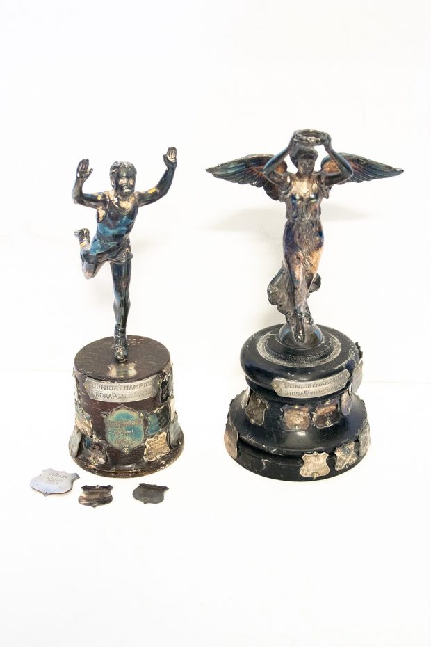 Deux petits trophées d’argent sur des bases en bois avec des écussons argent gravés.  Celui à gauche montre un homme en train de courir, les bras levés et tenant une couronne.  On voit trois écussons argent devant le trophée à gauche.