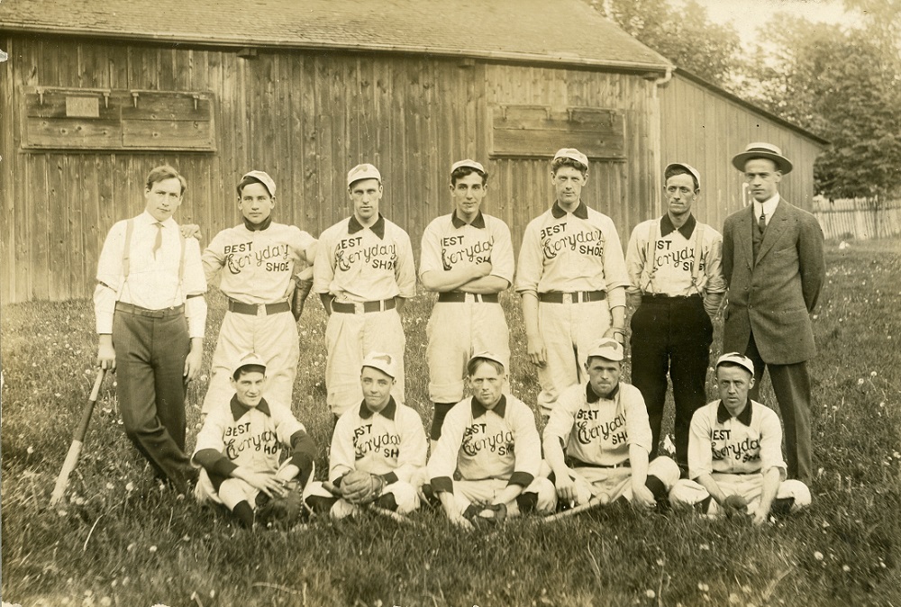 Photo en noir et blanc d’une équipe de joueurs de baseball sur deux rangées, le premier rang est assis sur l’herbe devant un bâtiment en bois ; Dix hommes en uniforme avec le logo « Best Everyday Shoe » sur leur jersey et deux hommes en costume de ville de chaque côté de la rangée arrière.