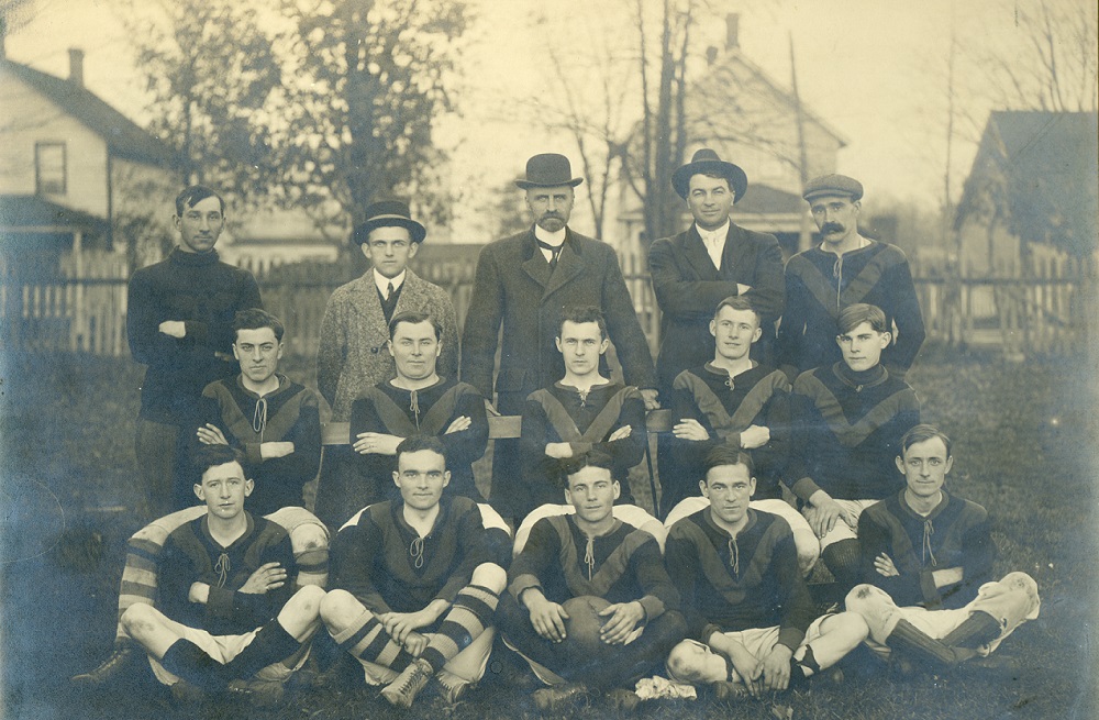 Photographie en noir et blanc d’une équipe de foot sur trois rangées, cinq joueurs sont en uniforme, l’un au milieu tient une balle, cinq joueurs sur la deuxième rangée sont assis sur un banc, sur la  dernière rangée, on voit  cinq joueurs debout avec au fond quatre maisons et une clôture en bois