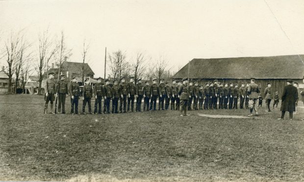 Photo en noir et blanc d’une rangée de jeunes gens en uniforme, portant un fusil.  Deux jeunes garçons et plusieurs hommes, dont certains en uniforme, regardent la scène.  A l’arrière-plan un grand bâtiment en bois et plusieurs maisons.