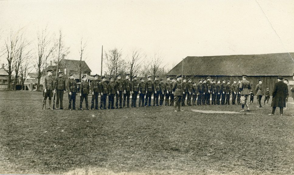 Photo en noir et blanc d’une rangée de jeunes gens en uniforme, portant un fusil.  Deux jeunes garçons et plusieurs hommes, dont certains en uniforme, regardent la scène.  A l’arrière-plan un grand bâtiment en bois et plusieurs maisons.