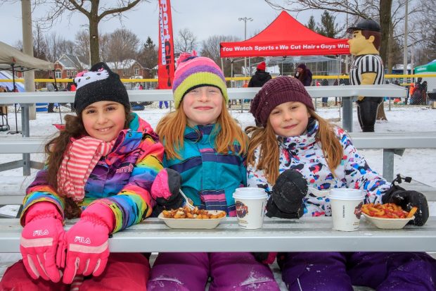 Photo en couleur de trois filles en costume de neige assises sur des gradins dans un parc ; deux des filles ont un bol de frites et des boissons.  On voit un personnage gonflable ( un arbitre) et des tentes à l’arrière.