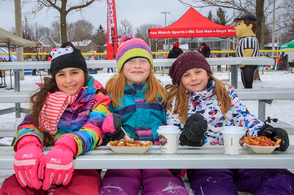 Photo en couleur de trois filles en costume de neige assises sur des gradins dans un parc ; deux des filles ont un bol de frites et des boissons.  On voit un personnage gonflable ( un arbitre) et des tentes à l’arrière.