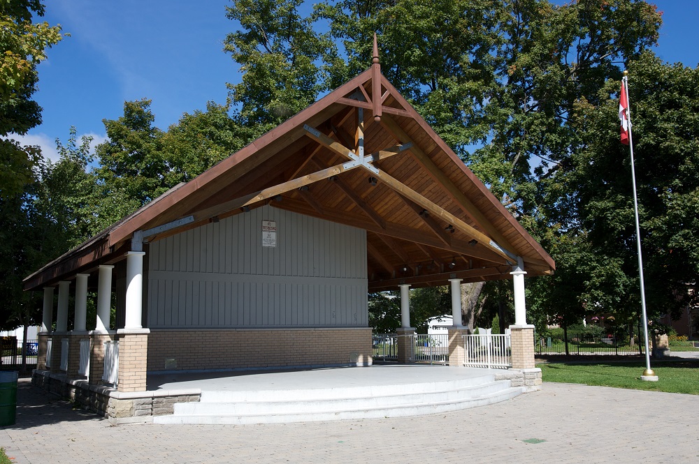 Photo en couleur d’un kiosque à musique couvert de forme carrée avec une estrade en ciment,  10 colonnes supportant un toit en bois et une structure en bois et en brique.  Il y a un drapeau canadien sur un poteau près du coin droit.  On voit à l’arrière de l’herbe, des arbres et une clôture.