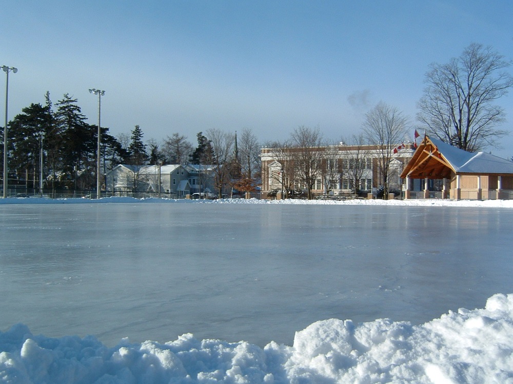 Photo en couleur d’une grande patinoire entourée de bancs de neige, différents bâtiments et terrain de baseball à l’arrière.