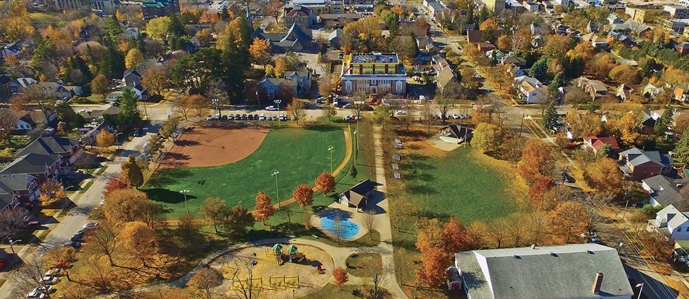 Photo couleur d’un parc en automne montrant un terrain de jeu et une fontaine à jets douchants dans le coin inférieur gauche, on voit une grand bâtiment blanc avec un toit gris dans le coin inférieur droit, un kiosque à musique couvert dans le coin supérieur droit et un terrain de baseball avec éclairage dans le coin supérieur gauche, des maisons et des bâtiments à l’arrière.