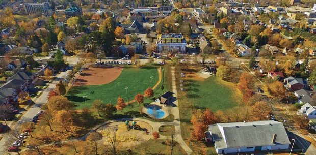 Photo couleur d’un parc en automne montrant un terrain de jeu et une fontaine à jets douchants dans le coin inférieur gauche, on voit une grand bâtiment blanc avec un toit gris dans le coin inférieur droit, un kiosque à musique couvert dans le coin supérieur droit et un terrain de baseball avec éclairage dans le coin supérieur gauche, des maisons et des bâtiments à l’arrière.