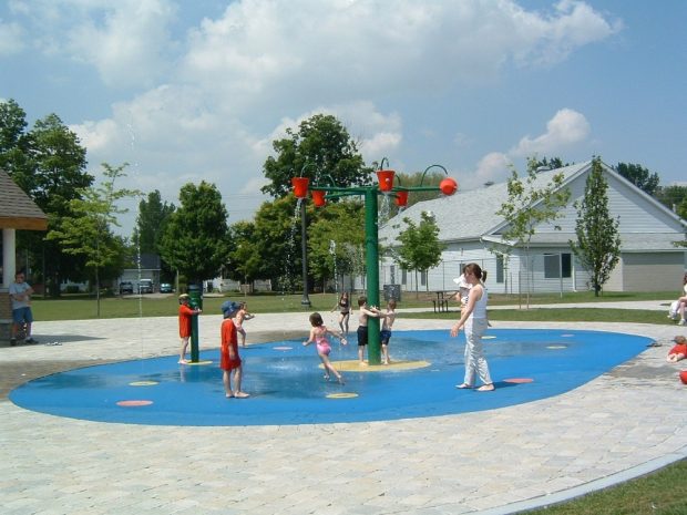 Photo en couleurs de sept jeunes enfants et d’une femme avec un bébé qui jouaient sous la fontaine.  On voit une surface ovale peinte en bleu et un poteau vert avec cinq seaux rouges qui se remplissaient d’eau ; 8 cercles rouges  avec des jets d’eau et une pompe verte.  Des pierres grises entouraient la surface, plusieurs personnes autour.  À l’arrière un grand bâtiment blanc en bois avec des arbres, de l’herbe et deux maisons.