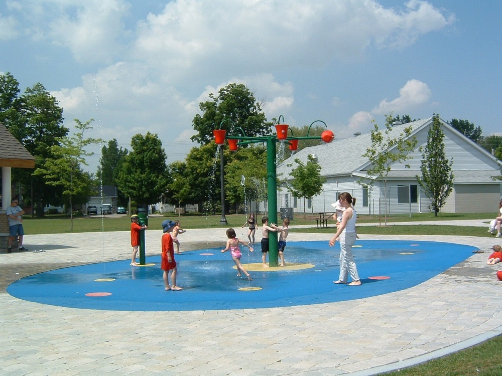 Photo en couleurs de sept jeunes enfants et d’une femme avec un bébé qui jouaient sous la fontaine. On voit une surface ovale peinte en bleu et un poteau vert avec cinq seaux rouges qui se remplissaient d’eau ; 8 cercles rouges avec des jets d’eau et une pompe verte. Des pierres grises entouraient la surface, plusieurs personnes autour. À l’arrière un grand bâtiment blanc en bois avec des arbres, de l’herbe et deux maisons.