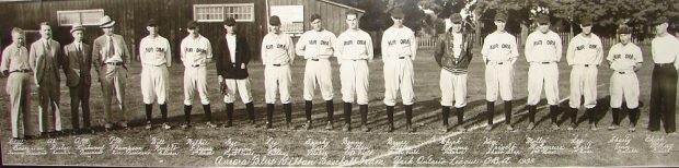 Photo panoramique en noir et blanc de 17 hommes alignés sur une pelouse, tous sauf quatre  à gauche portent un uniforme avec le nom de chaque joueur, juste en dessous.  À l’arrière des arbres et un bâtiment en bois.