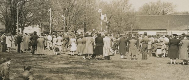 Photo un peu floue en noir et blanc d’un grand rassemblement de gens, beaucoup tournant le dos à l’appareil photo.  Douze personnes dans un parc  sont assises sur une estrade à gauche, quatre poteaux arborent  l’Union Jack et on voit des arbres et des maisons à l’arrière.