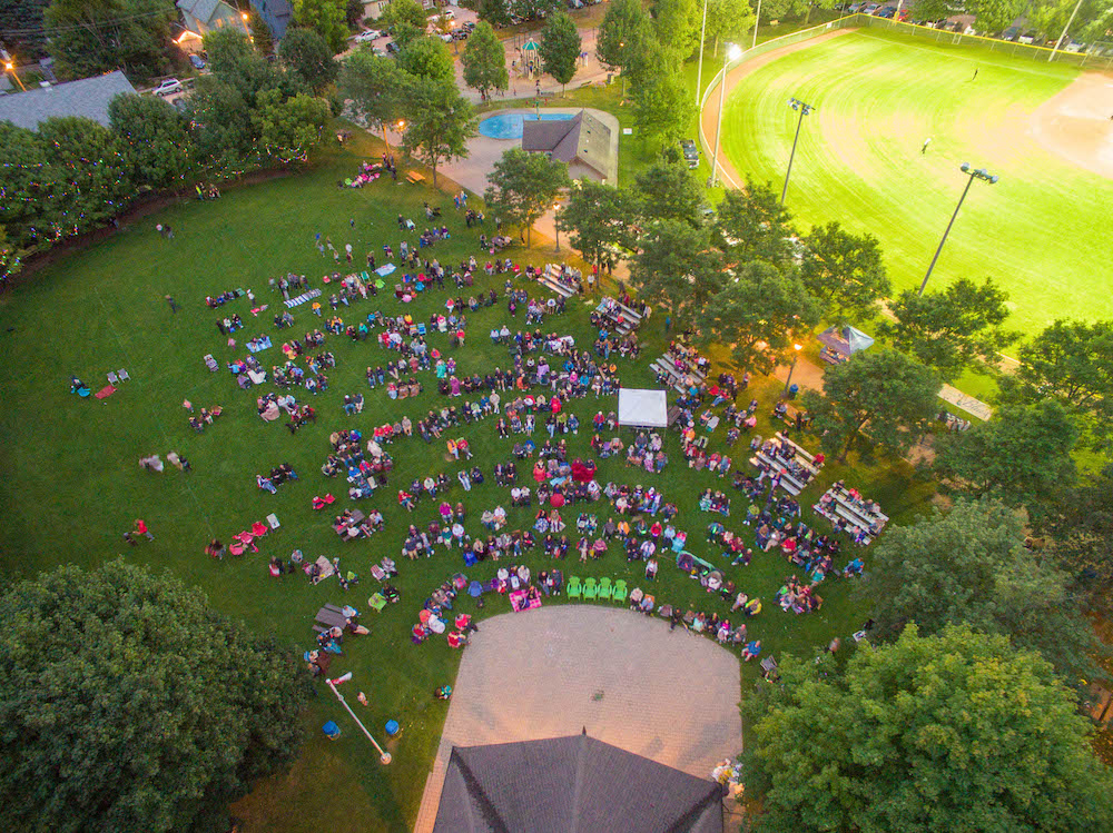 Photo en couleur prise par un drone qui montre un grand parc avec une scène couverte, une fontaine avec des jets douchants et un terrain de baseball.  La pelouse est remplie de gens qui regardent la scène assis sur des chaises ou des bancs.