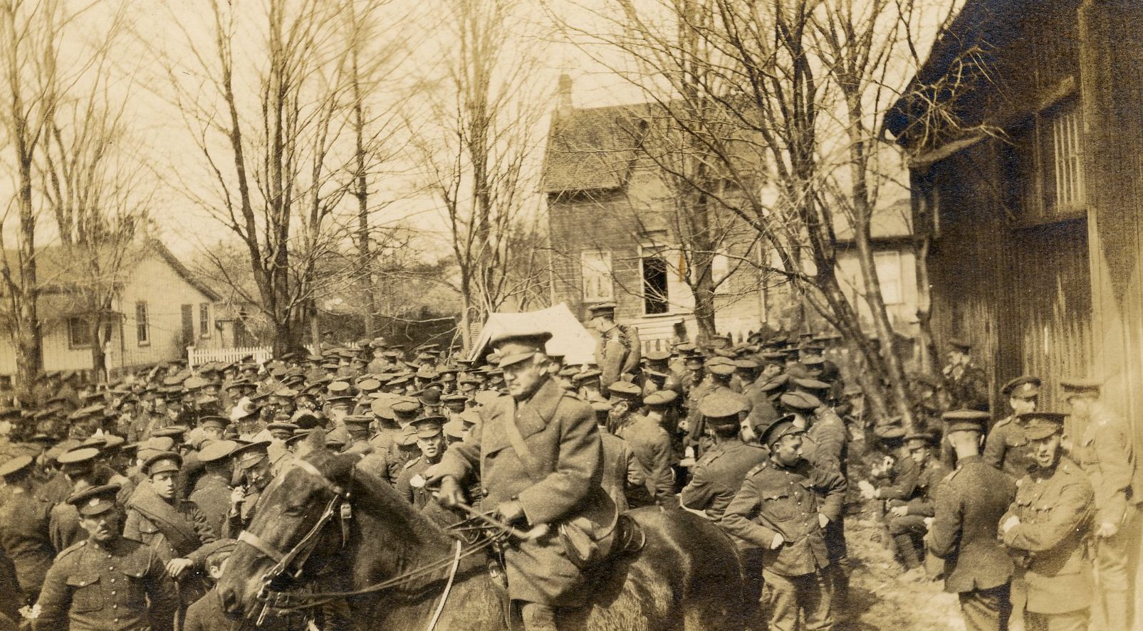 Photo en noir et blanc d’un large groupe de soldats à gauche d’un bâtiment en bois.  Officier à cheval et arbres et maisons à l’arrière.