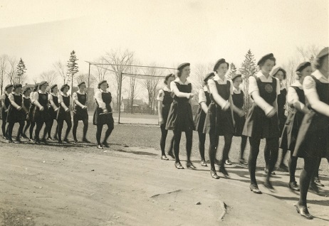 Photo en noir et blanc montrant des filles en uniforme défilant dans un parc.  Au fond, terrain de baseball et maisons