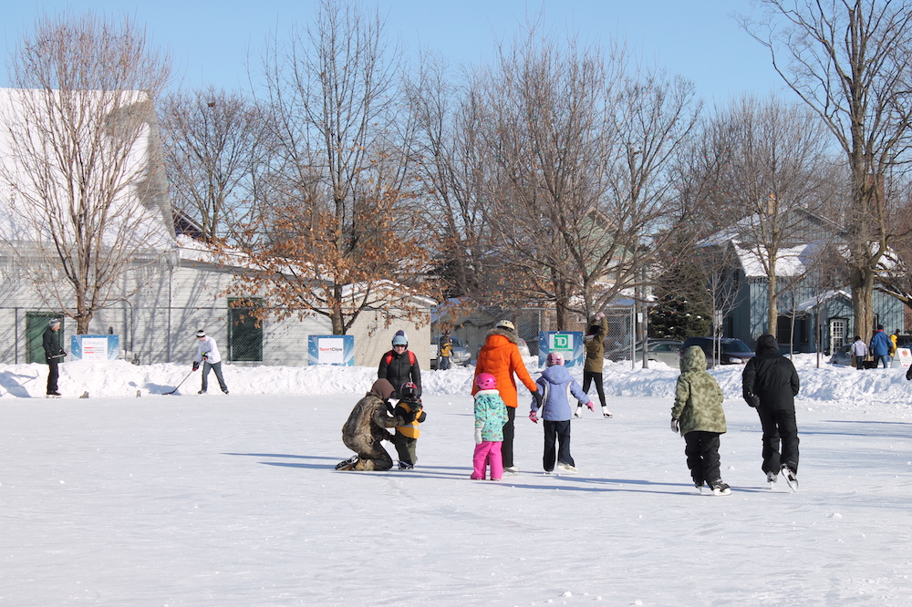 Photo en couleur prise en hiver montrant des gens de tous les âges patinant sur la patinoire en plein air.  Deux patineurs tiennent un bâton de hockey, à  gauche une clôture devant un  grand bâtiment, deux maisons à droite.