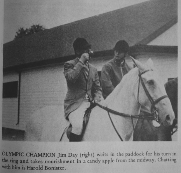 Photo en noir et blanc de deux cavaliers.  Ils portent une  bombe noire et les vêtements d’équitation de rigueur à l’époque.  L’homme à droite mange une pomme d’amour.  A l’arrière un grand bâtiment et une légende sous l’image.