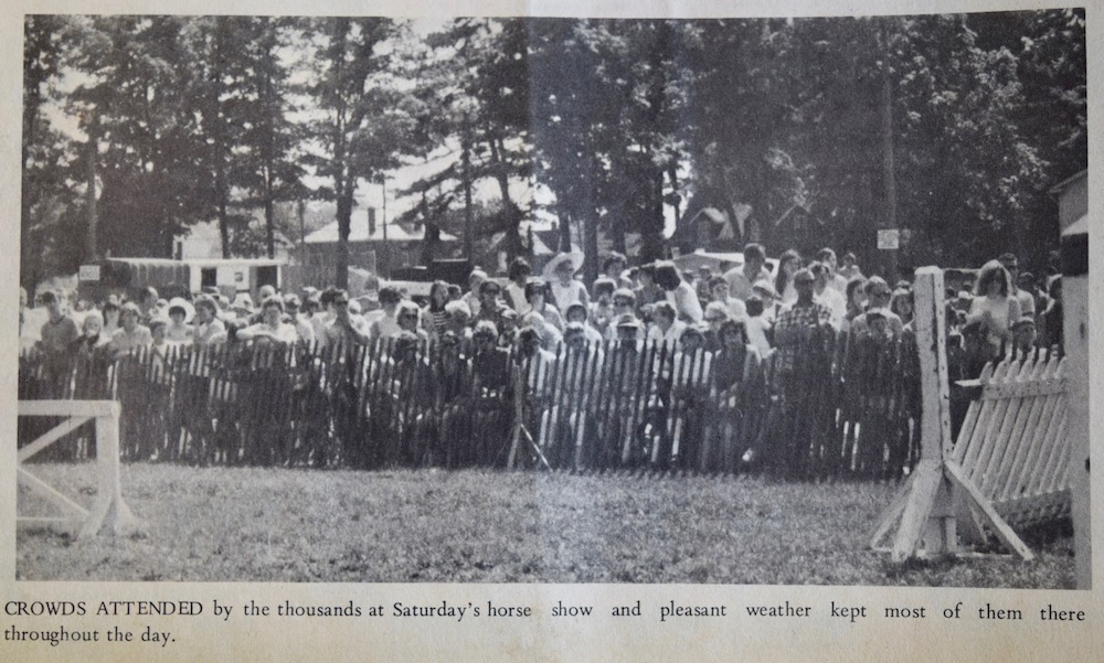 Image en noir et blanc avec une foule nombreuse dans le lointain derrière une clôture en bois.  Derrière de grands arbres et des maisons.  Devant, des structures en bois à droite et à gauche.  Légende sous l’image.