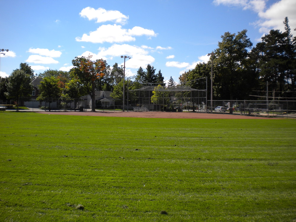 Photo en couleur du champ extérieur d’un terrain de baseball du côté du marbre.  On voit surtout la pelouse du champ extérieur, la terre rougeâtre du champ intérieur et une clôture derrière.