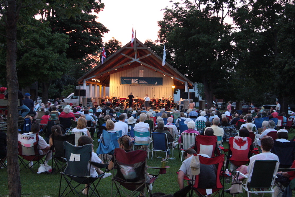 Photo en couleur de gens assis devant des musiciens qui jouent dans l’abri d’orchestre au crépuscule.