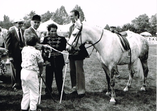 Photo en noir et blanc de 5 personnes et d’un cheval dans un parc, arbres et tentes au fond.