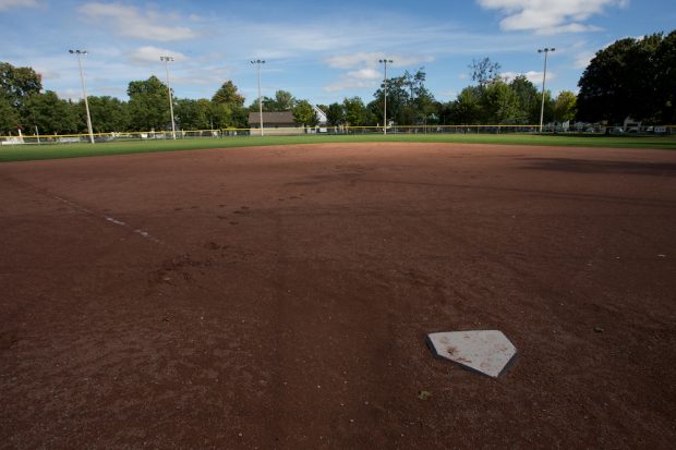 Photo couleur  d'un terrain de baseball prise derrière le marbre.  On voit principalement la terre rouge du terrain de baseball avec derrière la pelouse du champ extérieur bordé par une clôture.  On distingue à l'arrière une rangée de grands arbres et un petit bâtiment près du centre de l'image.