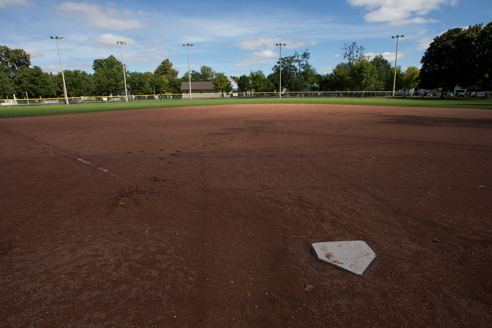 Photo couleur  d'un terrain de baseball prise derrière le marbre.  On voit principalement la terre rouge du terrain de baseball avec derrière la pelouse du champ extérieur bordé par une clôture.  On distingue à l'arrière une rangée de grands arbres et un petit bâtiment près du centre de l'image.