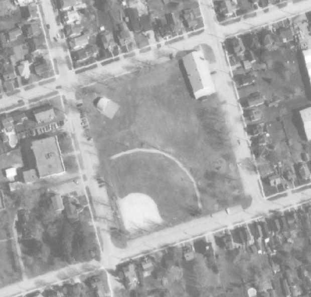 Photo aérienne en noir et blanc montrant un terrain de baseball, un abri d’orchestre et un manège militaire ainsi que les rues et maisons avoisinantes.