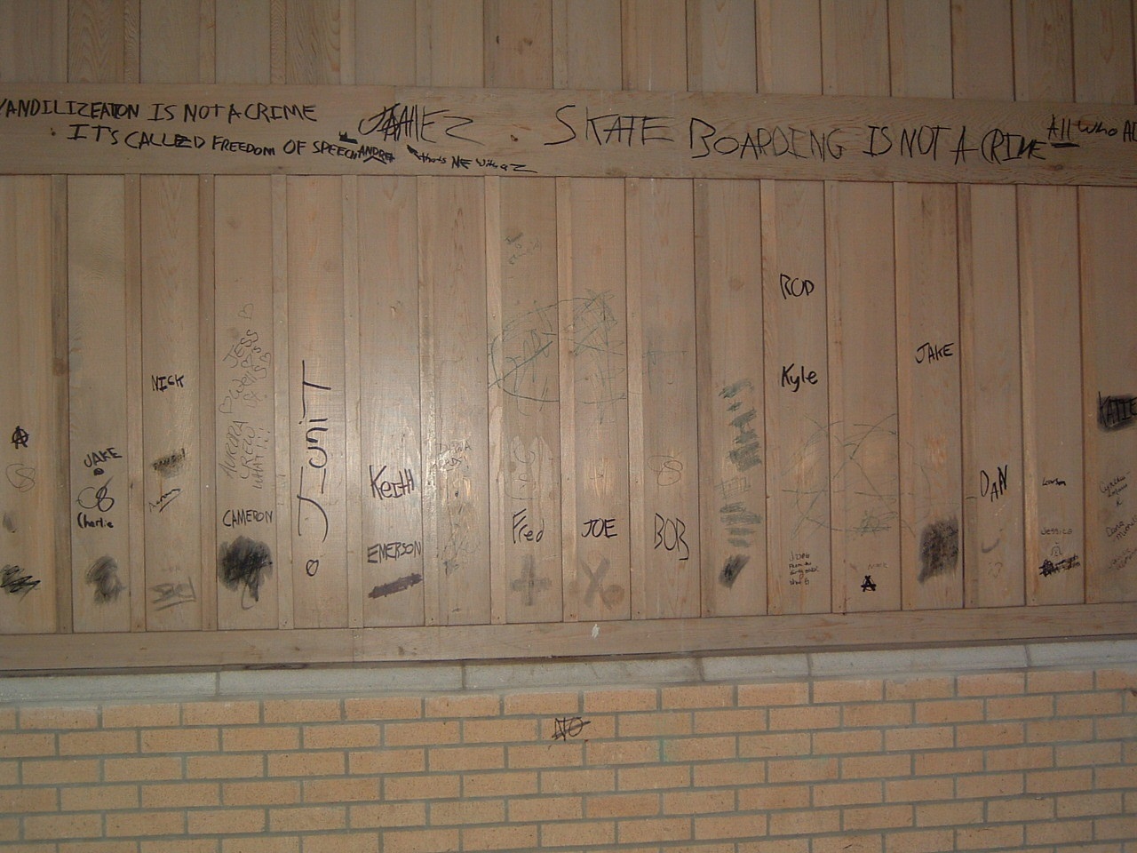 Photographie d’un mur en bois et en brique avec des graffiti à l’encre noire.
