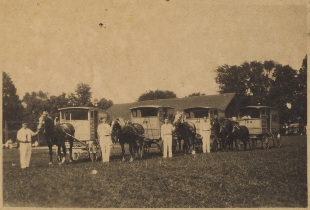 Une photo sépia des années 30 montre 4 hommes vêtus de blanc dans un parc,  A leur droite, un cheval tirant une carriole.  L’homme à gauche semble avoir quelque chose à la main.  Il y a un cinquième homme dans une carriole à l’extrême droite.  Au fond, on distingue un grand toit et la clôture d’un terrain de baseball.