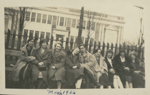 Une photo sépia datant de 1926 montre 9 filles assises sur un banc, une clôture en bois derrière elles et un grand bâtiment de deux étages au fond.  La photo a été prise de biais, la clôture et le bâtiment semblent pencher vers la droite.