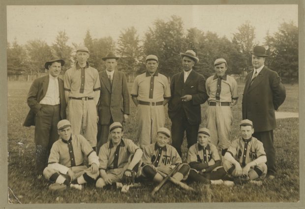Image sépia de sept hommes debout et cinq assis devant, posant pour une photo de l'équipe.  Ils sont dans un parc et huit d'entre eux portent un uniforme