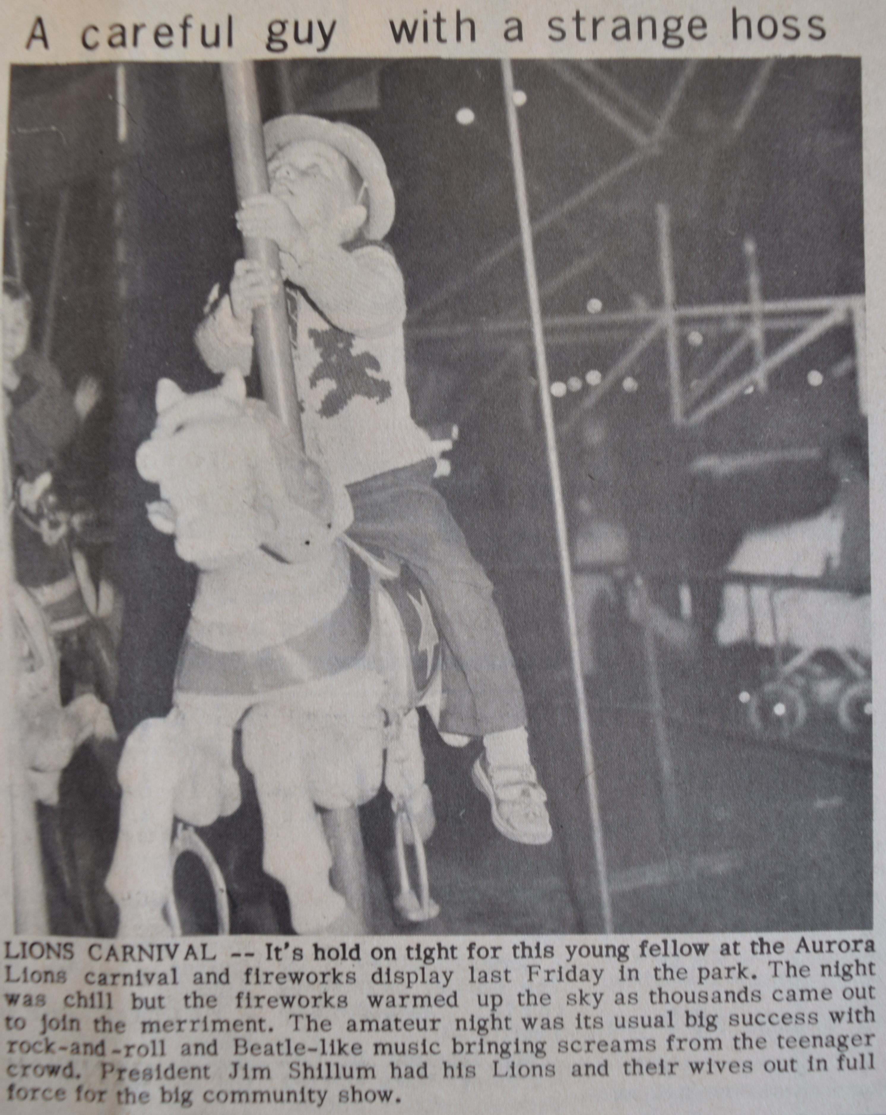 On voit une coupure de journal avec une  photo en noir et blanc montrant un petit garçon monté sur un cheval de manège, il porte un chapeau et regarde le ciel, au fond , un landau, des gens et une grande roue.
