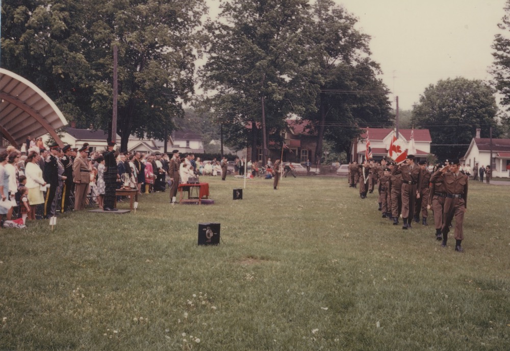 Photo en couleur avec des cadets qui défilent dans un parc à droite et se dirigent vers le photographe.  À gauche des gens les regardent.  On voit le toit de l’abri d’orchestre des maisons et de grands arbres.