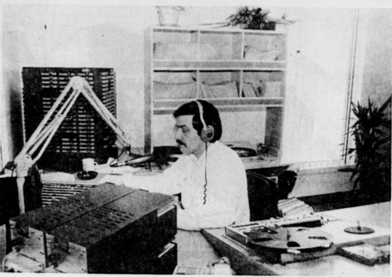 Photographie en noir et blanc de Michel animant une émission. Il a un micro devant lui et les écouteurs sur la tête. Il est entouré d’un équipement radiophonique.