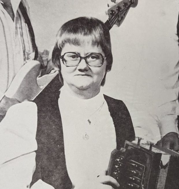 La photographie montre Francine Desjardins assise avec un accordéon sur les genoux et entourée de son groupe. Derrière elle, on peut remarquer un musicien avec une guitare basse.