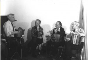 Photographie en noir et blanc, Philippe Bruneau assis sur un banc de piano, un accordéon à la main.  Alfred Couillard, Lorenzo Picard et Joseph Messervier le regarder jouer. Plusieurs micros sont déposés devant eux.