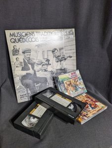 Photographie couleur montrant un vinyle, dont la pochette montre deux violonistes, deux cassettes VHS (Video Home System) avec des étiquettes de couleur jaune et une écriture manuscrite, deux disques compacts aux couleurs flamboyantes.