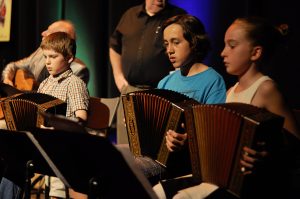 Photographie de trois jeunes accordéonistes en concert. Devant eux, des lutrins tiennent leur partition. Leur professeur, debout derrière, les surveille.