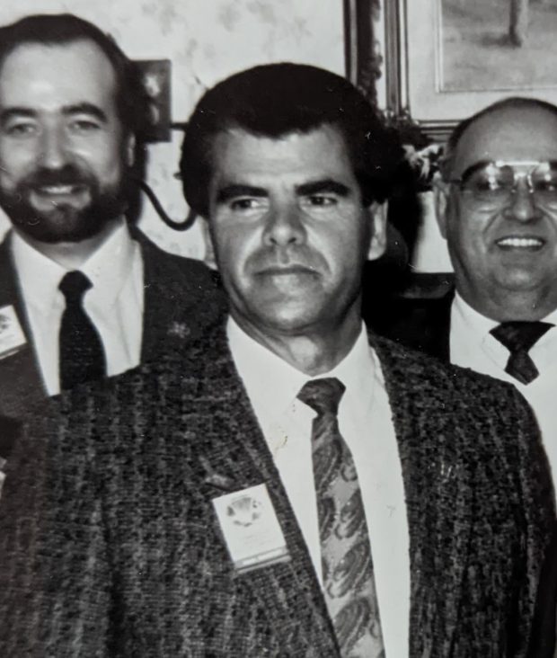 Photographie en noir et blanc de Marcel Messervier. Il est vêtu d'un veston raillé et d'une cravate à motif. Deux personnes en complet et cravate se tiennent debout derrière lui.