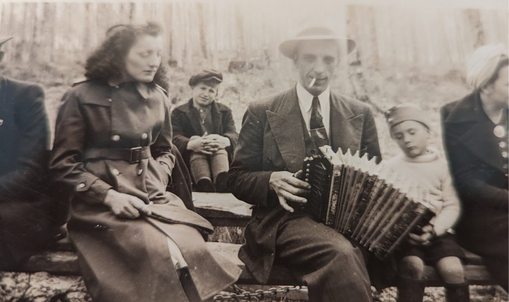 Photographie en noir et blanc de l’accordéoniste Fernand Fraser durant une partie de sucre jouant avec son accordéon sur les genoux. Une femme est assise auprès de lui et, derrière eux, un enfant.