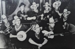 Black and white photograph of the Orchestre Montminy. Front row, left to right: Gérard Nicole, Gérard Montminy, Patrice Gaudreau, Maurice Gaudreau and Joseph Montminy. Second row, left to right: Conrad Belley, Ulric Côté, Réal Montminy and Camille Nicole. The musicians are all wearing cowboy hats and neckerchiefs. They are posing in front of a false backdrop.