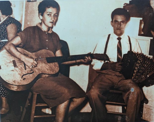 Photographie sépia montrant le couple et duo formé par Yvette Coulombe à la guitare et Ulric Lacombe à l’accordéon. Assis sur des chaises, ils jouent lors d’une veillée de danse familiale.