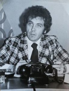 Photographie en noir et blanc montrant Gilles Vallière, une pipe et un crayon à la main, assis derrière un bureau. Devant lui se trouve un téléphone.